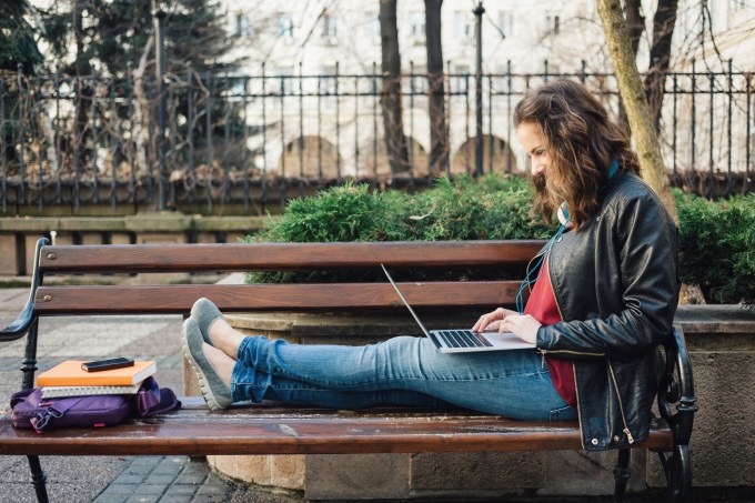 Estudante digitando em um laptop e estudando no banco de um jardim