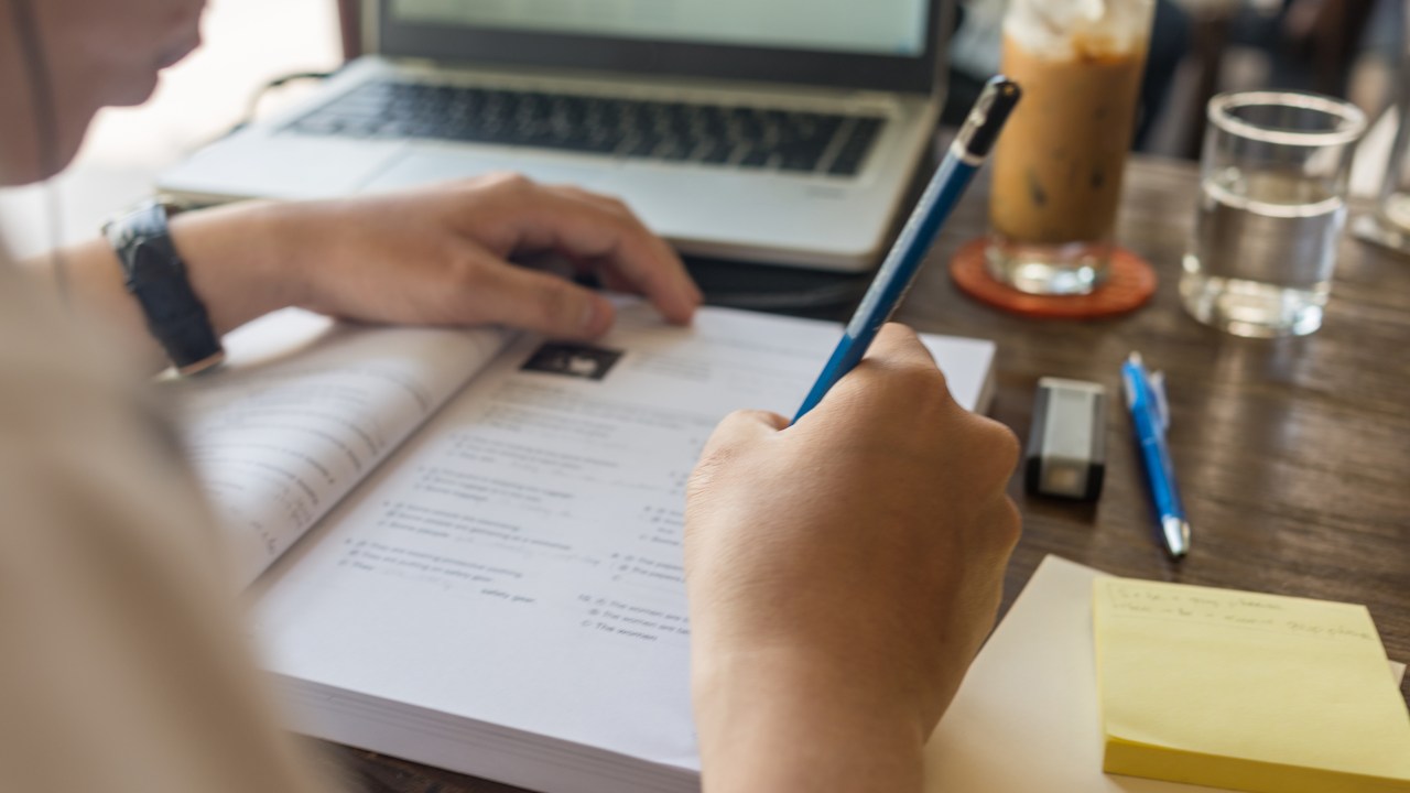 Estudante escrevendo com um lápis em um livro. Em cima da mesa, ao lado, estão um computador, materiais escolares e copos com água e café