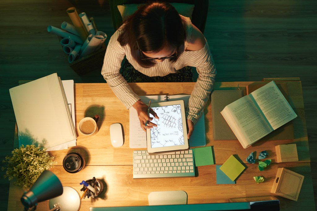 Uma aluna estuda sobre uma mesa de madeira. Na sua frente estão vários objetos de estudo, como livros, canetas, post-its e o teclado de um computador. Ela também segura um iPad.