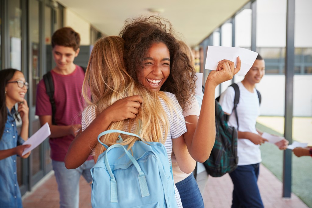 Duas garotas se abraçam e comemoram a aprovação no vestibular. Uma das jovens está de costas para a câmera. A outra de frente é é possível ver o rosto dela sobre o ombro da colega, enquanto ela observa o papel de aprovação