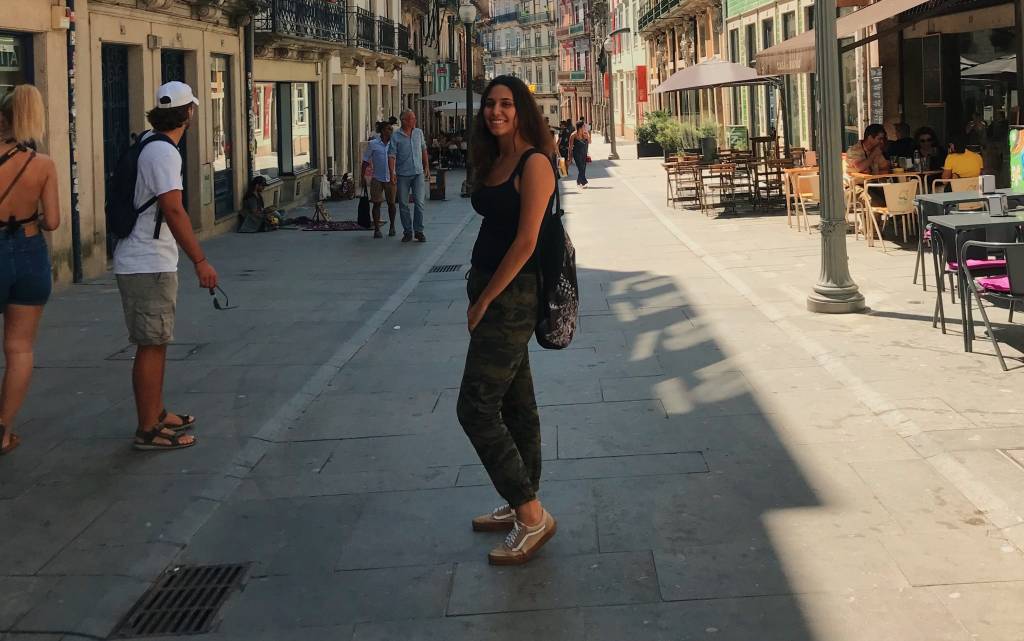 Estudando em Portugal 1 Mulher posa no meio da rua, em centro turístico de Portugal. Com um semblante feliz, ela usa roupas pretas e uma mochila. Na calçada, pessoas passam.