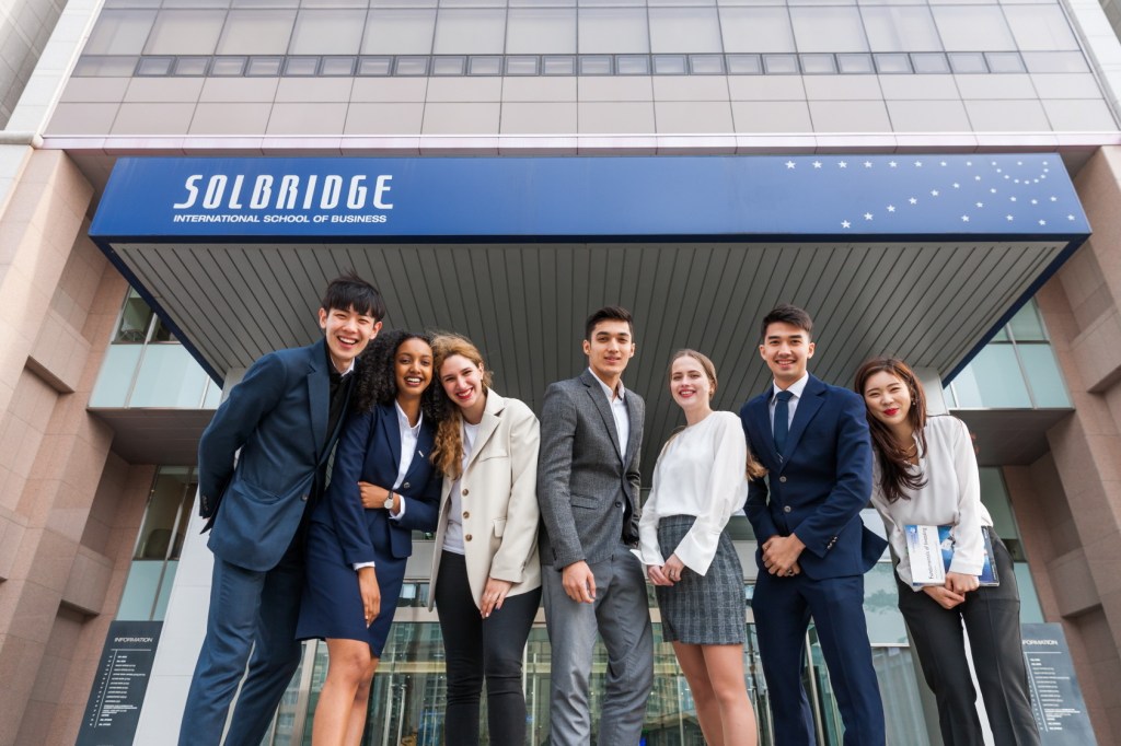 Grupo de 8 jovens, usando roupas empresariais, posa junto sorrindo em frente a um prédio da universidade, onde vemos o escrito "SolBridge: International School of Business"