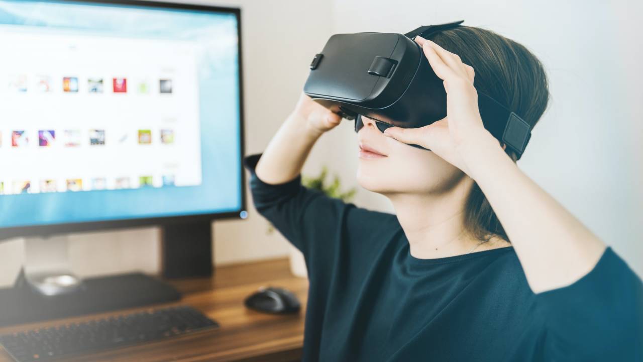 Jovem branco de cabelos castanhos curtos usa camiseta preta enquanto coloca as duas mãos em um óculos de Realdiade Virtual que está em seu rosto. Atrás dele, há um computador ligado em uma mesa de madeira.
