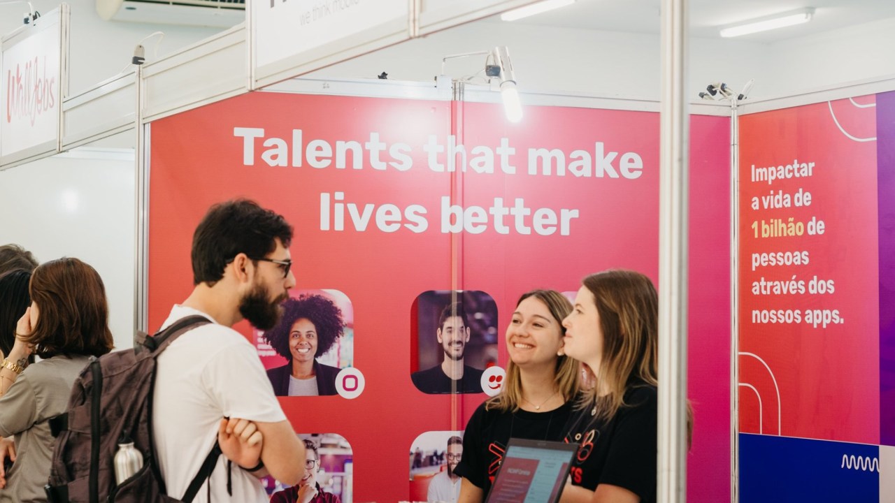 Foto com um homem branco de cabelo negro e barba em uma stand vermelha com duas mulheres de preto sorrindo para ele. Há na parede escrito "talents that make lives better"