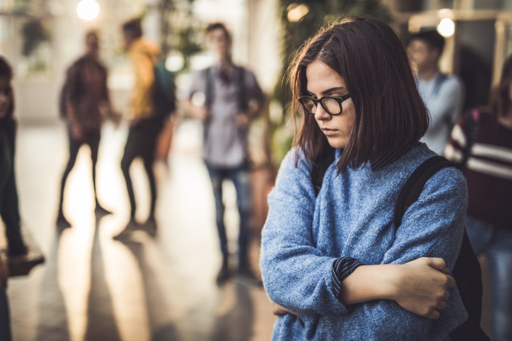 No primeiro plano, uma garota cabisbaixa. E no fundo, estudantes conversando