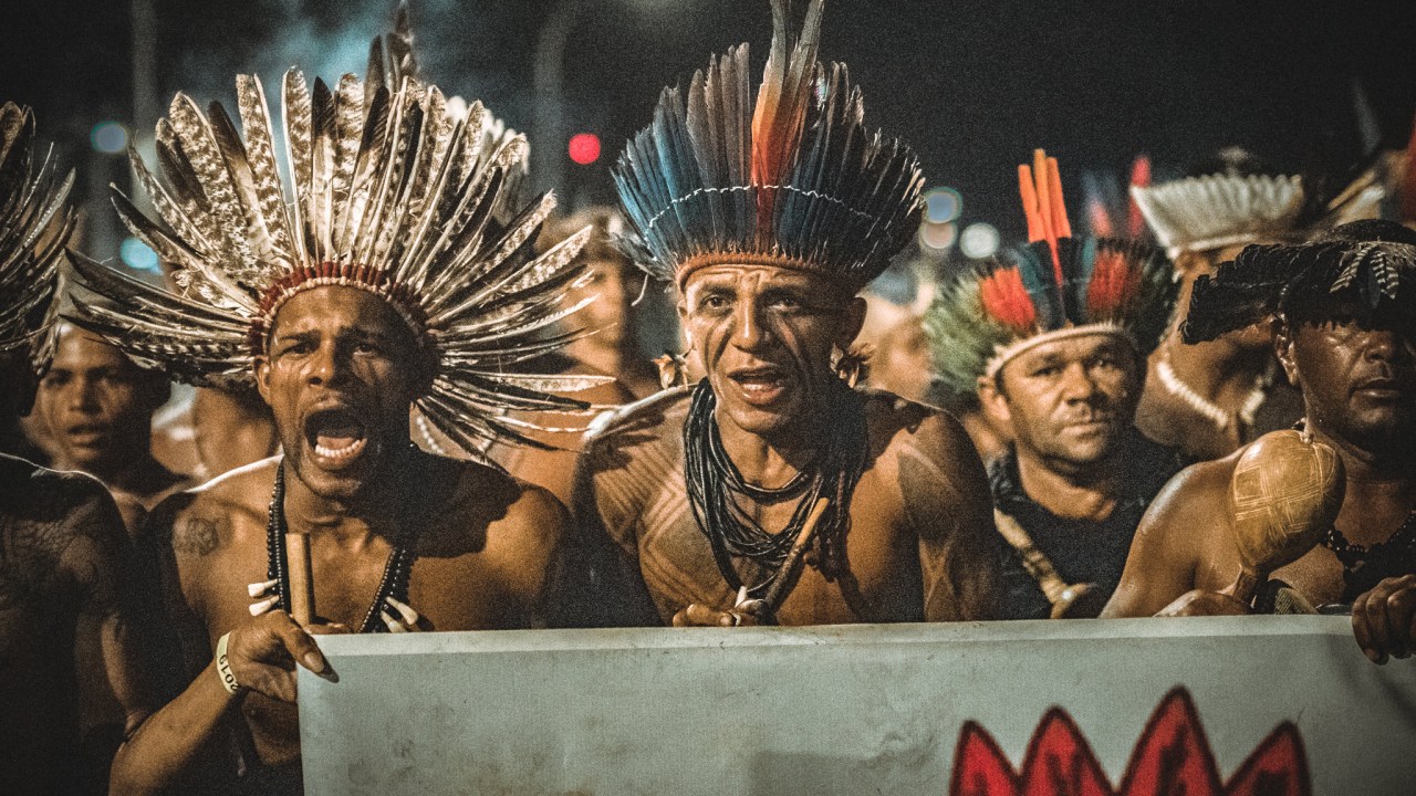 A foto foca em três homens indígenas segurando um cartaz. Atrás deles, há outras pessoas no que parece ser um protesto. Está de noite.