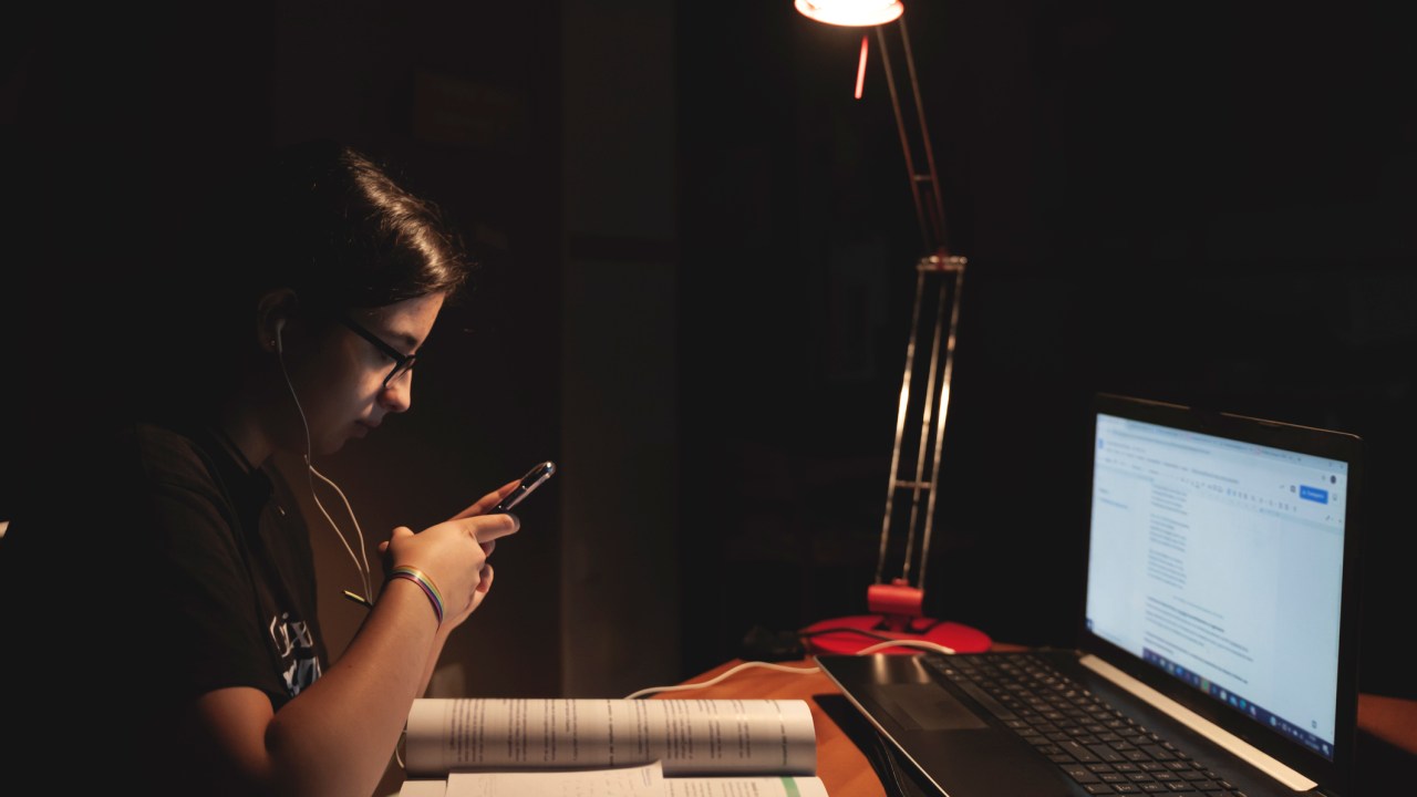 Estudante está em um quarto com notebook, apostilas, celular e fone de ouvido. Mesmo com uma lâmpada, o quarto está escuro.