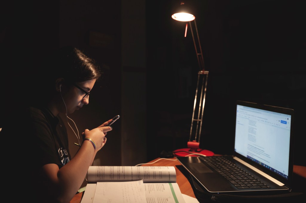 Estudante está em um quarto com notebook, apostilas, celular e fone de ouvido. Mesmo com uma lâmpada, o quarto está escuro.