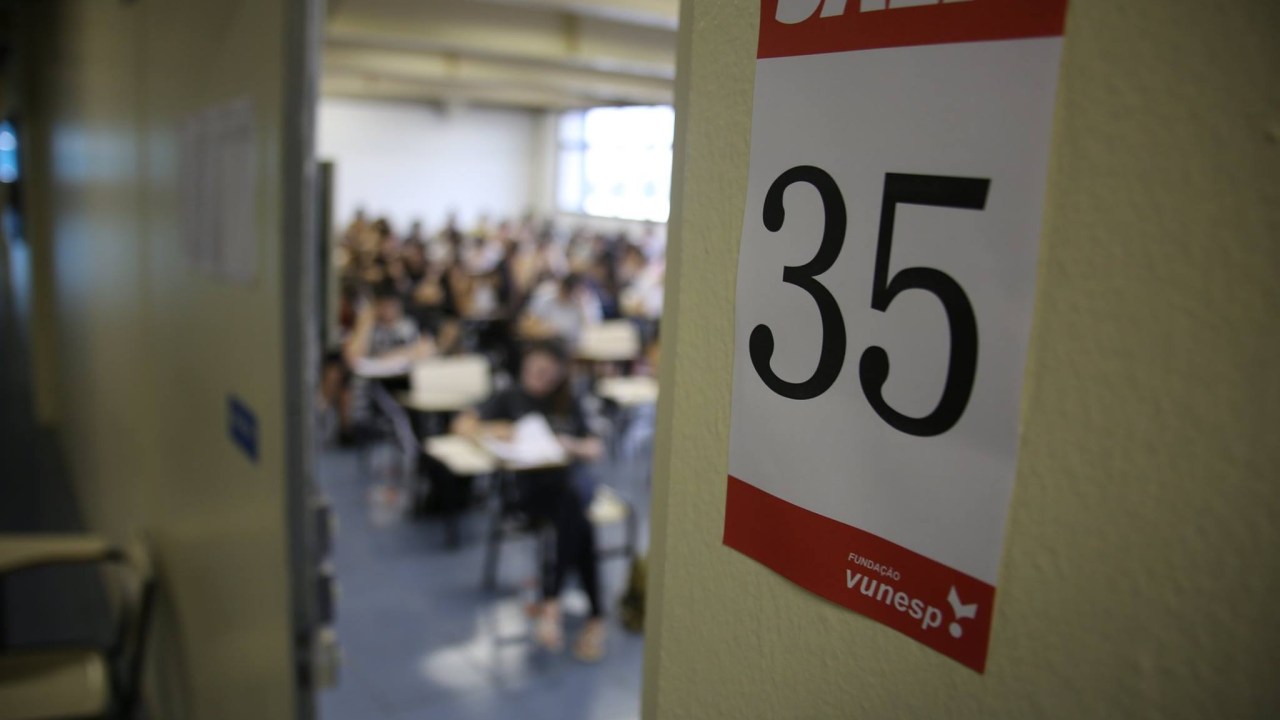 Sala de aula de aplicação de prova vestibular. A fotografia mostra uma sala de aula por meio de uma porta entre aberta, na porta está colado um papel escrito "Sala 35". Dentro da sala, se vê estudantes sentados em carteiras realizando uma prova.