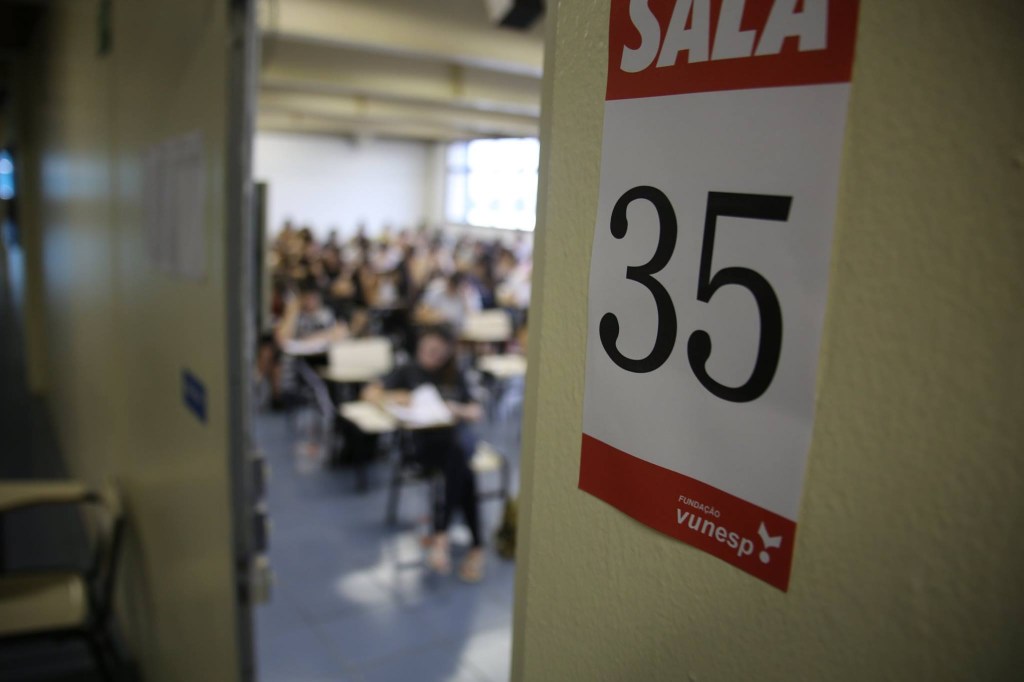 Sala de aula de aplicação de prova vestibular. A fotografia mostra uma sala de aula por meio de uma porta entre aberta, na porta está colado um papel escrito "Sala 35". Dentro da sala, se vê estudantes sentados em carteiras realizando uma prova.