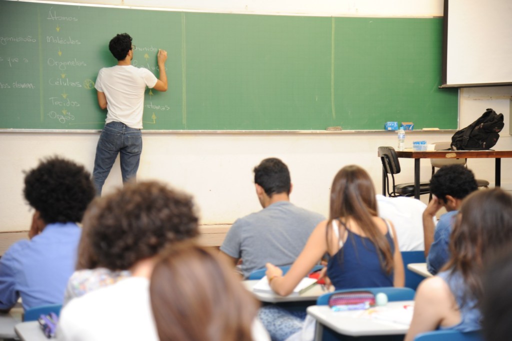 Quatro alunos dentro da sala de aula. Eles estão sentados assistindo a aula que um professor está dando.