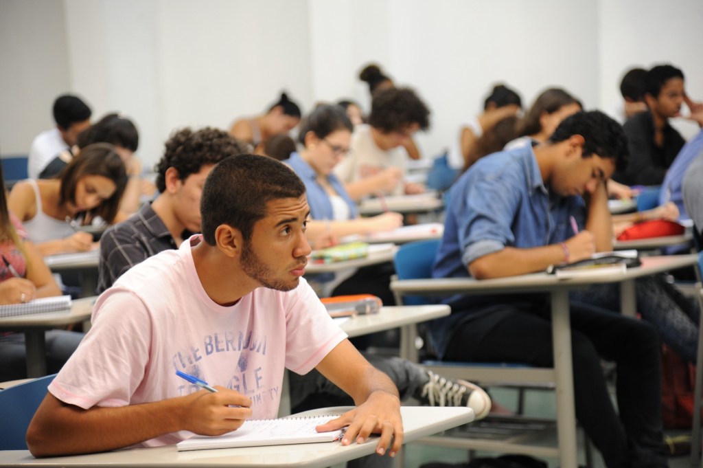 Sala de aula de um cursinho com vários estudantes sentados prestando atenção nas aulas.