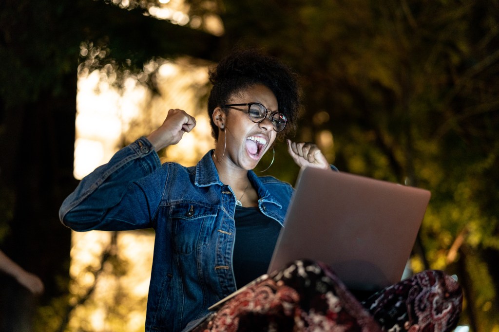 Jovem negra vestindo jaqueta jeans comemora aprovação em frente ao computador