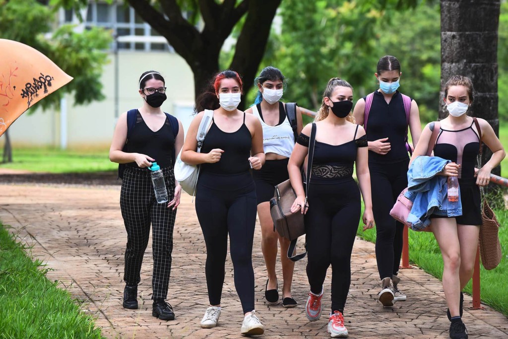 Grupo de jovens mulheres máscara. Algumas estão usando roupas esportivas, e outras estão com bolsas, casacos e garrafa d'água na mão