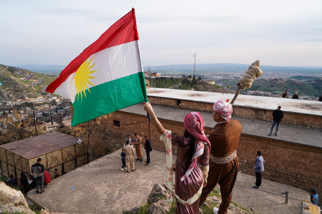 Na imagem, um homem e uma mulher curdos erguem uma bandeira do Curdistão ao lado de uma igreja