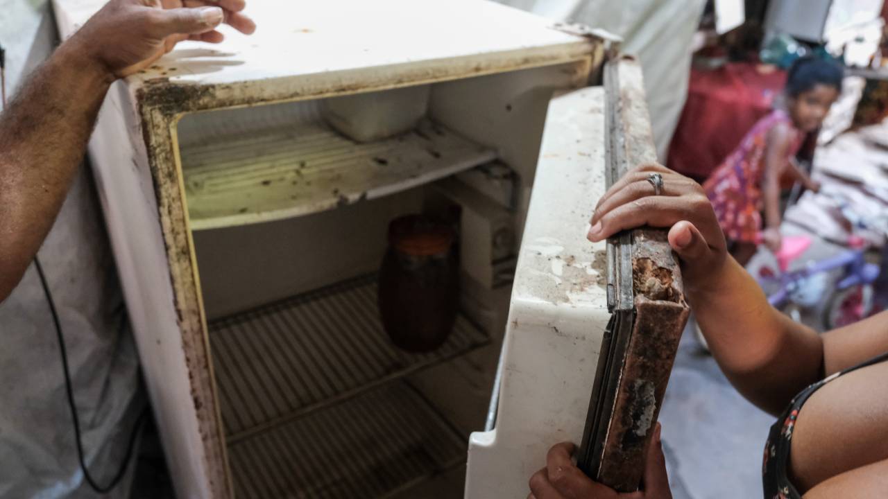 Mulher mostra geladeira vazia. Criança ao fundo observa a cena