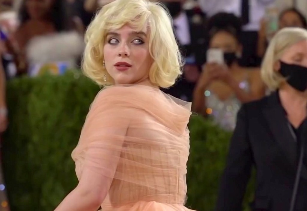 Billie Eilish A cantora Billie Eilish tem pele branca clara, cabelos loiros acima dos ombros, olhos azuis e usa um vestido salmão.