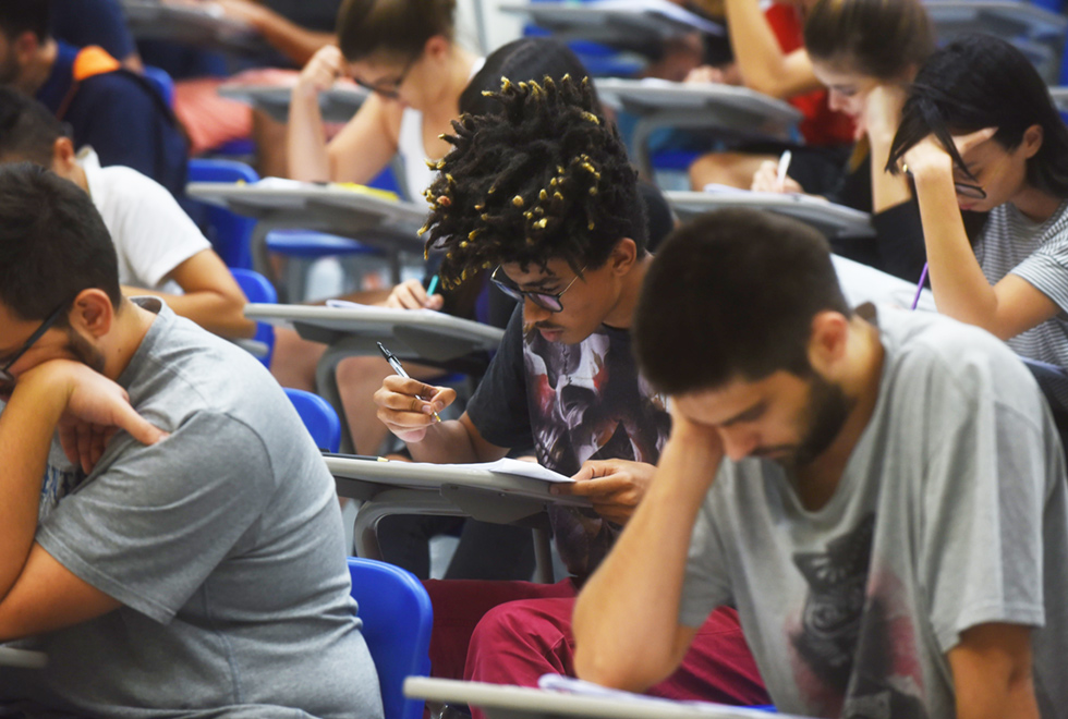 Fotografia mostra parte de uma sala de aula, de aplicação de prova. Jovens estão sentados em carteiras, olhando concentrados para uma prova, com uma caneta nas mãos. O foco da imagem é um jovem negro, na casa dos seus 20 anos, de camisa estampada, que está escrevendo em sua prova.