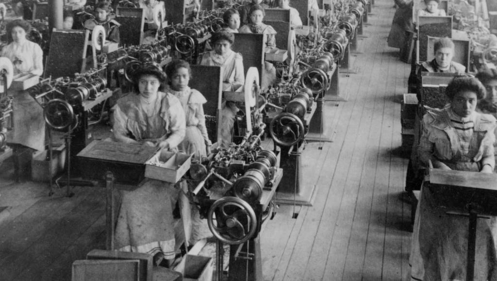 Mulheres-trabalhando-fábricas-século-20 Mulheres trabalhando operando máquinas, elas estão sentadas e olham para a câmera.