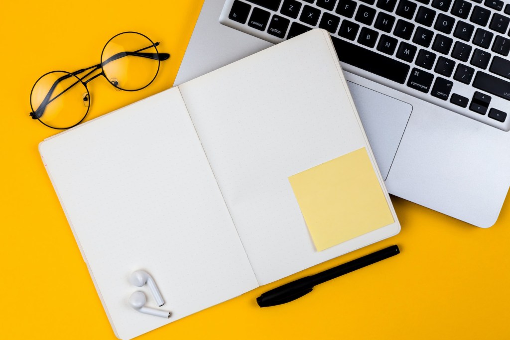 Fotografia mostrar visão superior de uma mesa de cor amarelo vivo, nela estão: um caderno aberto, com as folhas em branco, um óculos de grau, parte de um notebook e um parte de fones de ouvidos sem fio.