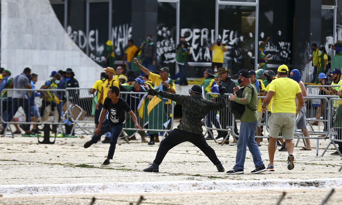 pessoas vestidos de preto ou de verde e amarelo destroem com pedras e outros objetos a sede dos três poderes. Na imagem, um homem atira uma pedra