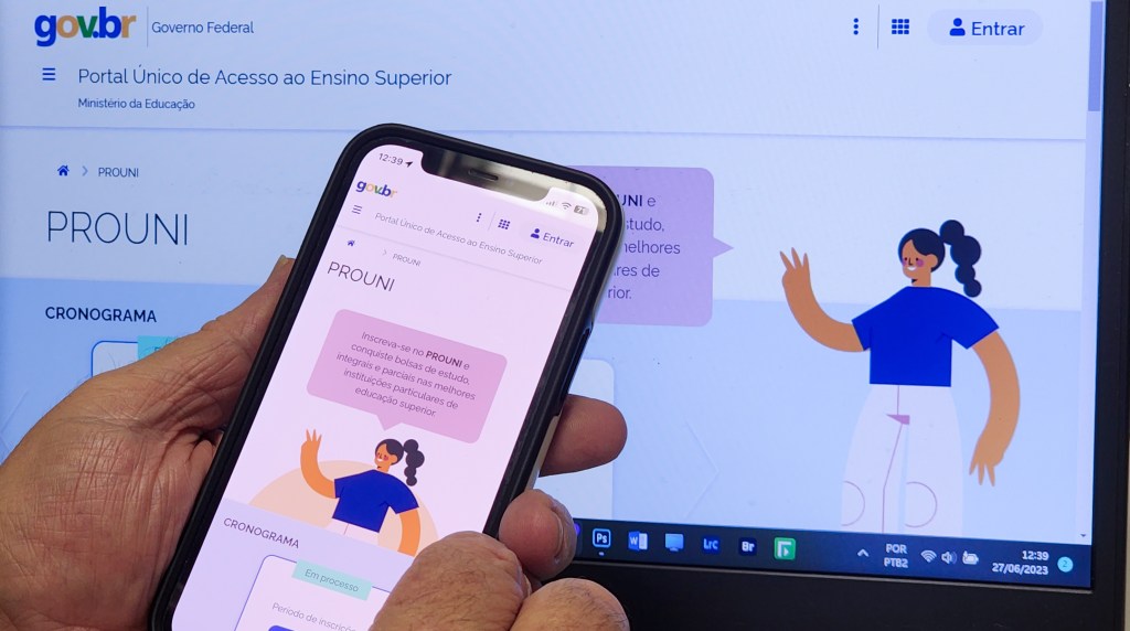 Mão segurando celular em frente à tela de comptuador com o site do Prouni aberto