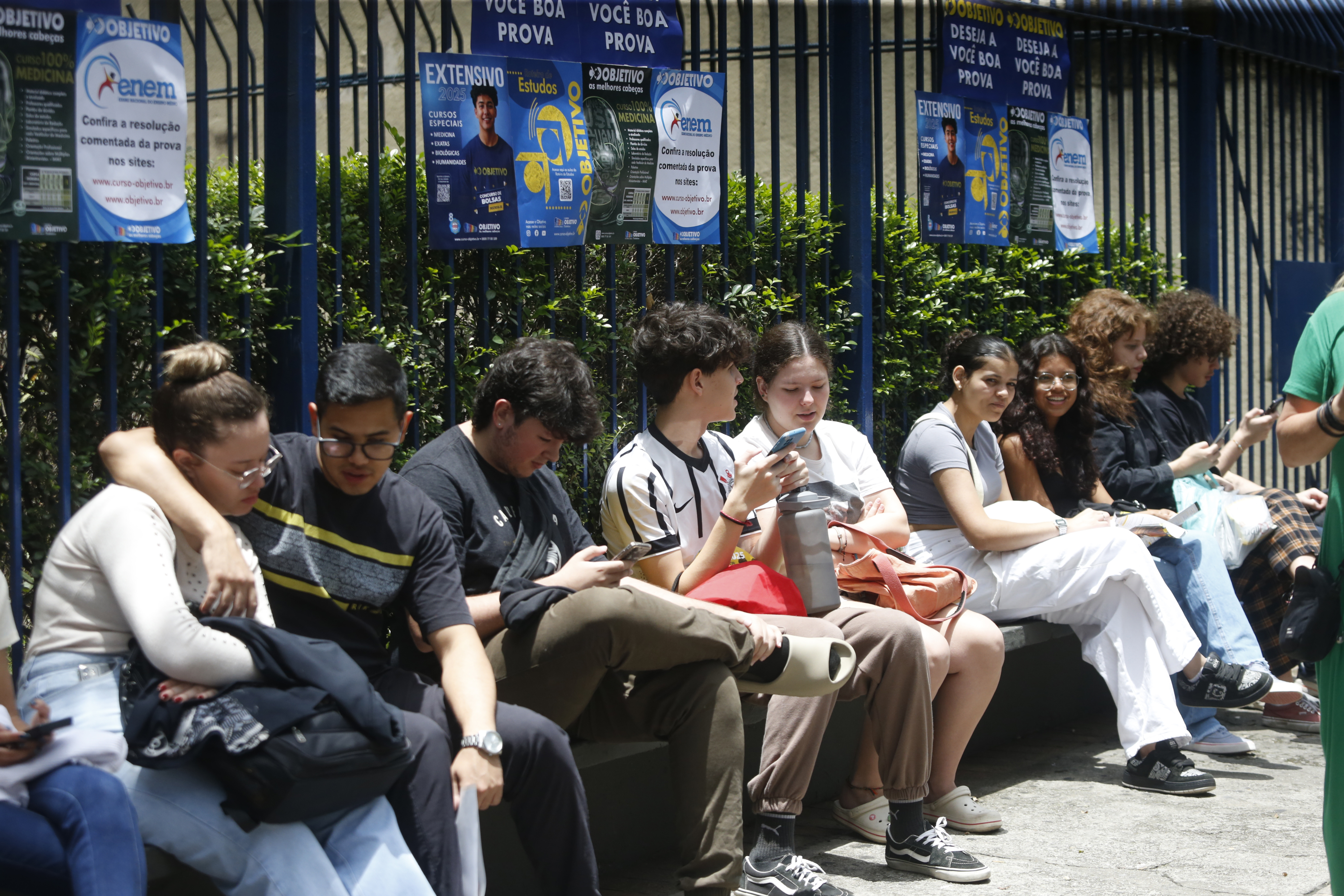 Candidatos do Enem aguardam em área externa em fila do local de prova, conversando e rindo.